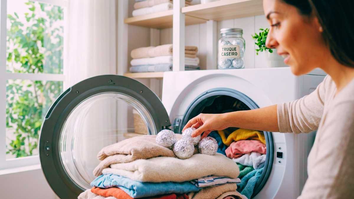 Fotografia autêntica de estilo de vida em plano médio-aproximado, mostrando uma mão feminina colocando três bolinhas compactas de papel alumínio (do tamanho de bolinhas de tênis) dentro do tambor de uma máquina de secar/lava e seca de carregamento frontal aberta. O tambor está cheio de roupas coloridas, fofas e levemente úmidas, como blusas de lã, toalhas e camisetas de algodão. O foco está nítido nas bolinhas de alumínio metálico e nas texturas dos tecidos. A cena é iluminada por luz natural suave vindo de uma janela à esquerda, que revela vegetação verde borrada ao fundo. O ambiente é uma lavanderia doméstica acolhedora e moderna, com prateleiras de madeira branca contendo toalhas dobradas, um pequeno vaso de planta verde e um jarro de vidro retrô com o rótulo escrito à mão 'TRUQUE CASEIRO' (com mais bolinhas de alumínio dentro) utilizados para secar roupa. A atmosfera é útil, otimista e curiosa, capturando a descoberta de um segredo doméstico eficiente. A foto tem profundidade de campo rasa e granulado natural de filme.