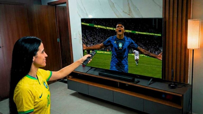 Mulher comemorando gol da seleção brasileira na TV em alta definição, garantida por uma antena digital interna posicionada no rack da sala moderna.
