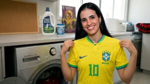 Home 13 Torcedora sorrindo na lavanderia, vestindo e segurando a camisa do Brasil número 10 da Seleção Brasileira, ao lado de uma máquina de lavar moderna, ilustrando o passo a passo de como lavar camisa de time sem danificar o tecido ou o silk.