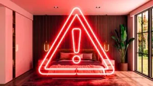 Imagem conceitual de um quarto luxuoso e moderno onde, flutuando sobre uma cama perfeitamente arrumada com roupa de cama 100 algodão branca e travesseiros macios, há um grande ícone de alerta de perigo (triângulo com exclamação) em neon vermelho brilhante. O brilho do neon reflete sutilmente sobre a roupa de cama branca.